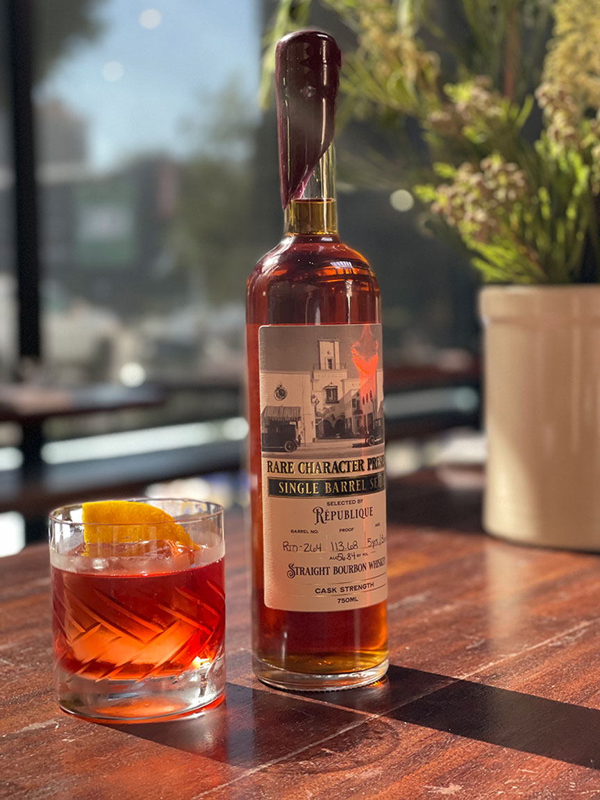 a boulevardier cocktail next to a bottle of republique's limited edition bourbon a top a wooden table in the restaurant