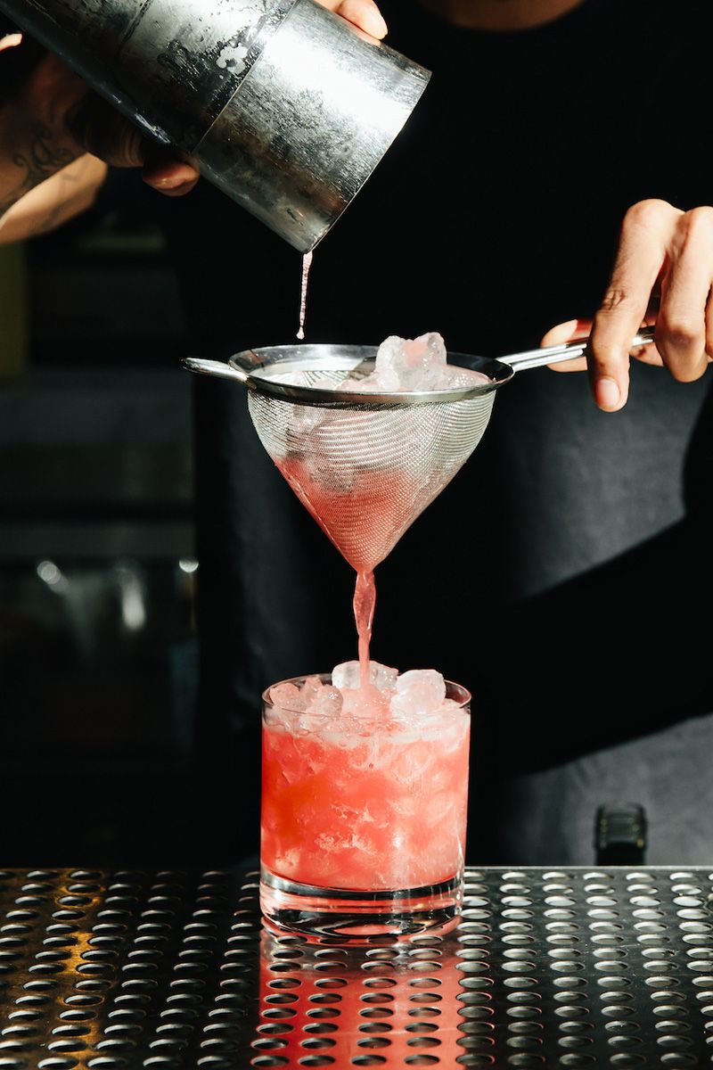 bartender pouring a pink cocktail out of a shaker, through a strainer, and into a rocks glass